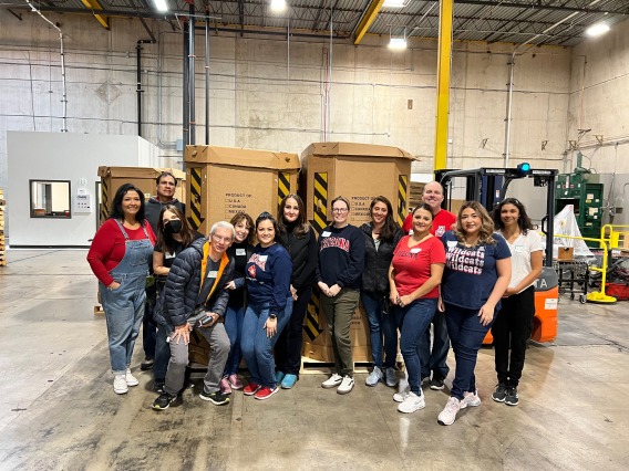 A group of UA Cares volunteers at the Community food bank. 13 people standing in front of cardboard boxes in a warehouse.