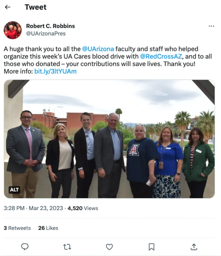 A knee-up photo of University of Arizona employees who volunteered to run the UA Cares Blood Drive on Tuesday, March 21, 2023, standing with President Robert Robbins. They are on an outside balcony in the Student Union building. Behind them are red brick buildings of the campus and a partly-cloudy sky. 